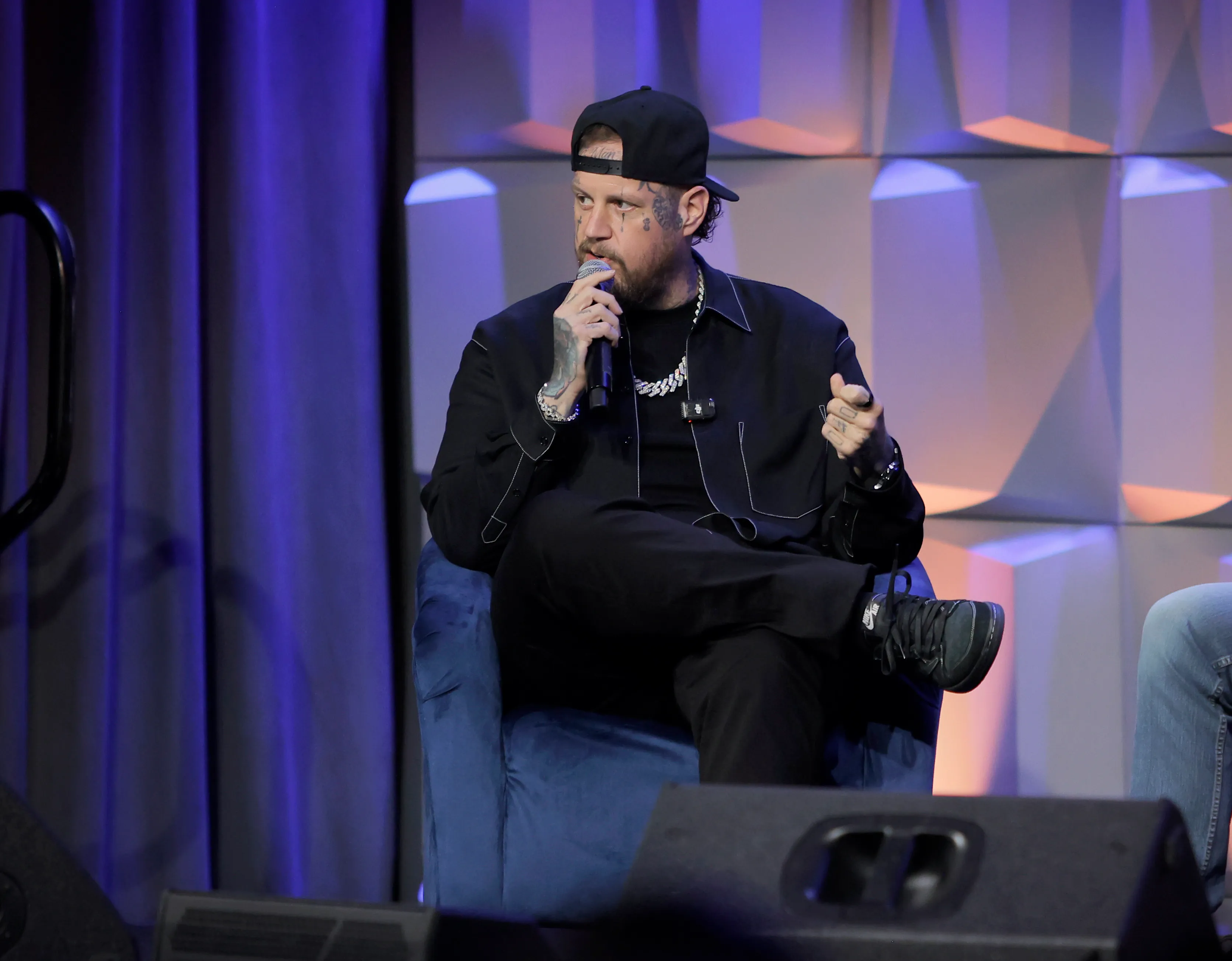 Jelly Roll speaking at an event, wearing a black shirt, black baseball cap, and holding a microphone.