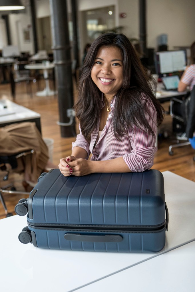 Jen Rubio, co-founder of Away Luggage, smiling with her hands resting on a blue Away suitcase.