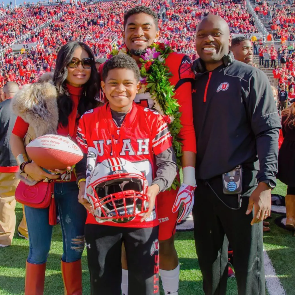 A family of four, including two young men in Utah Utes jerseys, poses on a football field in front of a stadium full of cheering fans.