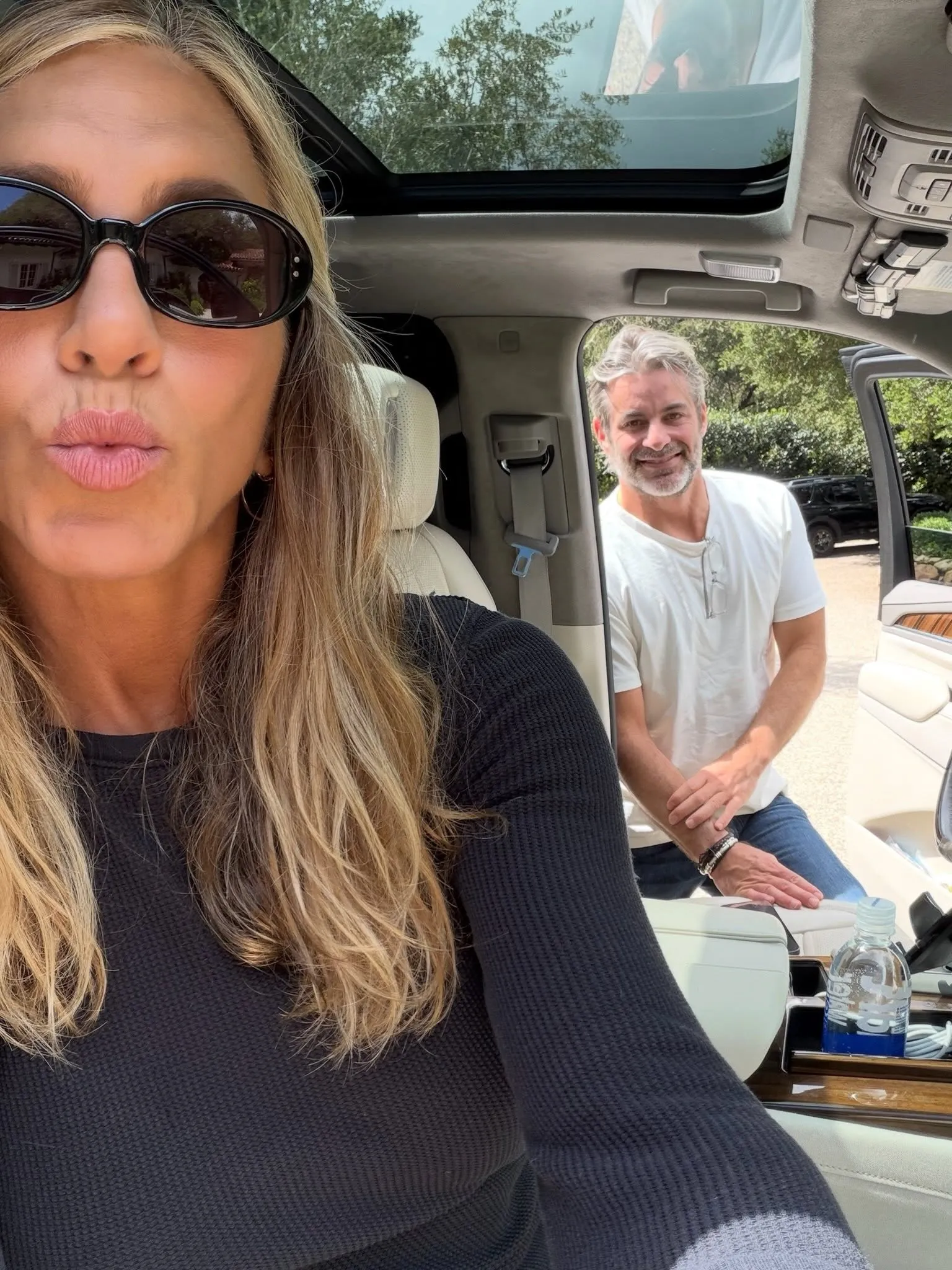 Jennifer Aniston in sunglasses makes a kiss face while a man smiles in the background, leaning against a car door.
