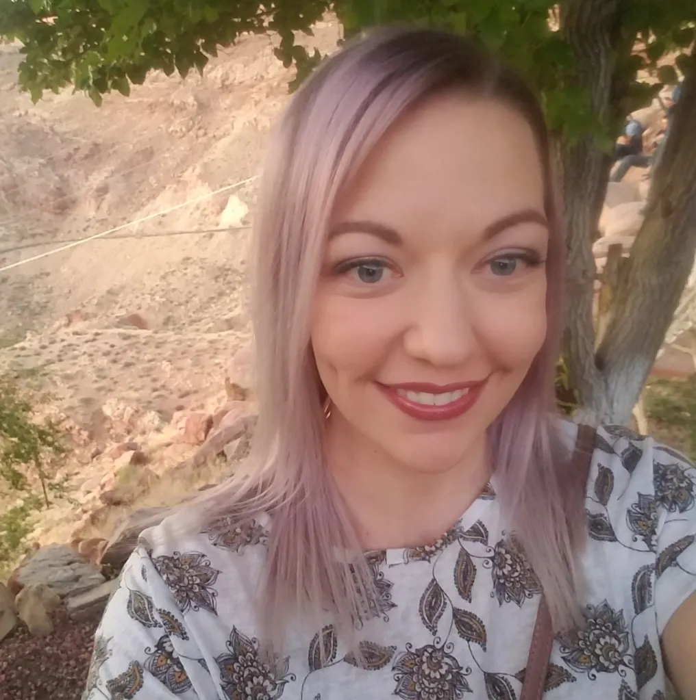 Jessica Orton Lyman, a woman with long light purple hair and a floral shirt, smiling in an outdoor setting with rocky hills and trees in the background.