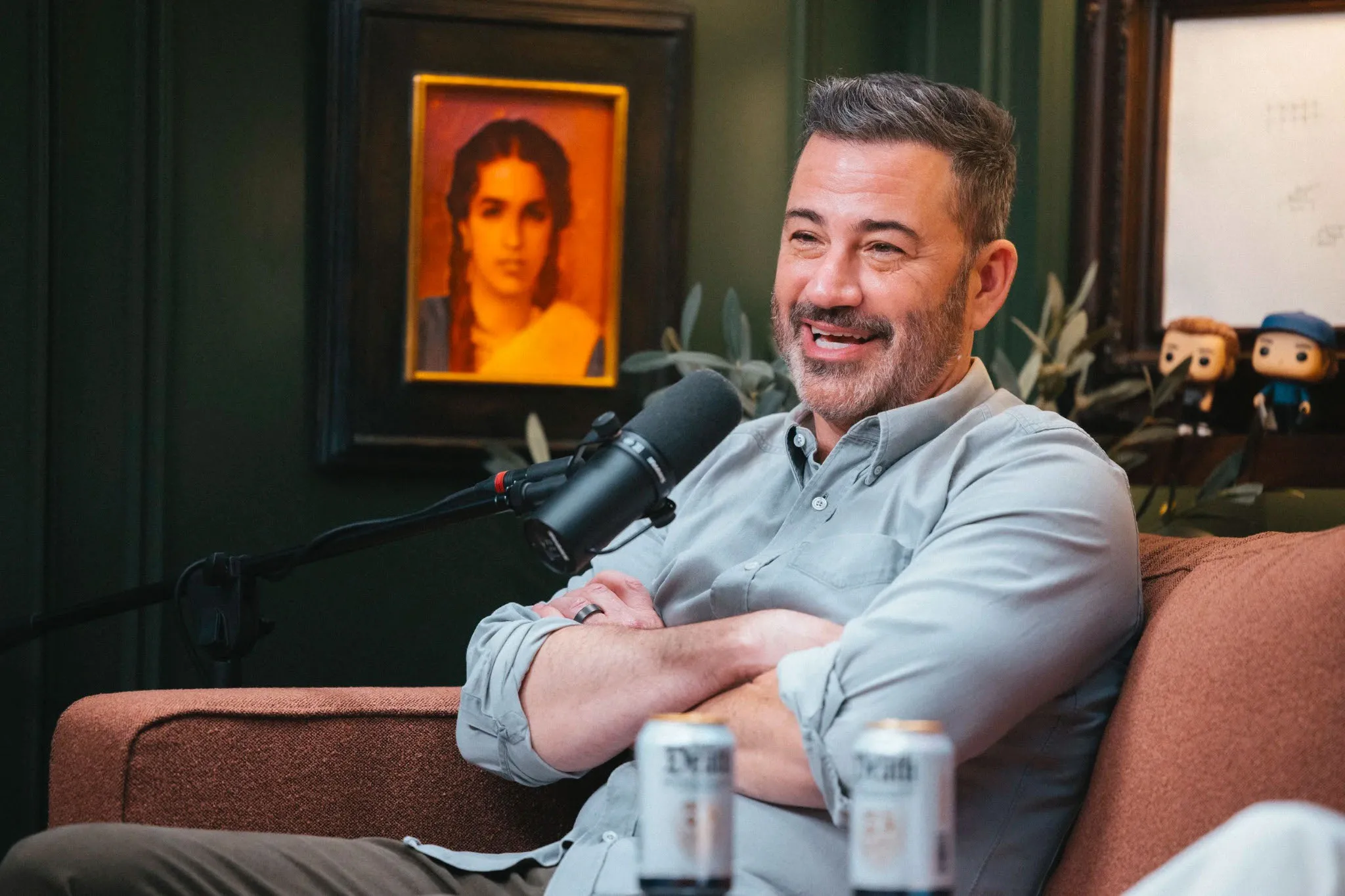 Jimmy Kimmel smiling while sitting on a brown couch with a microphone in front of him.