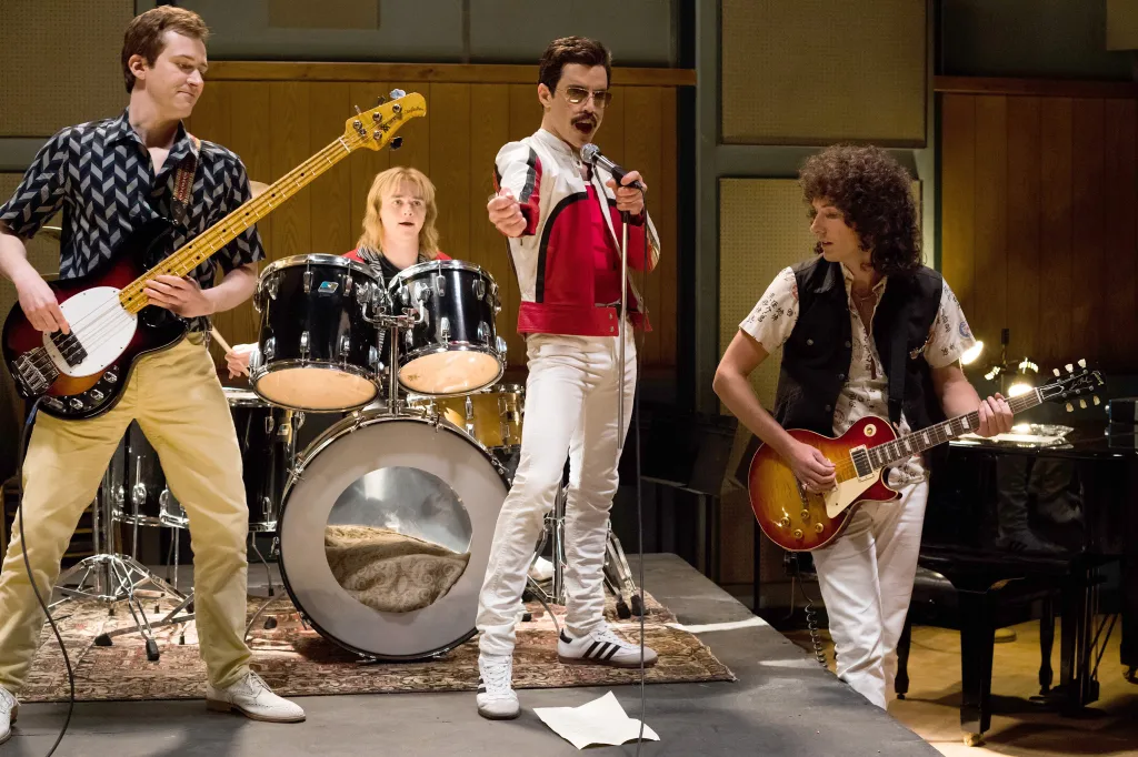 Four actors portraying the band Queen, with Freddie Mercury singing into a microphone, Brian May playing guitar, John Deacon playing bass, and Roger Taylor behind the drum kit.