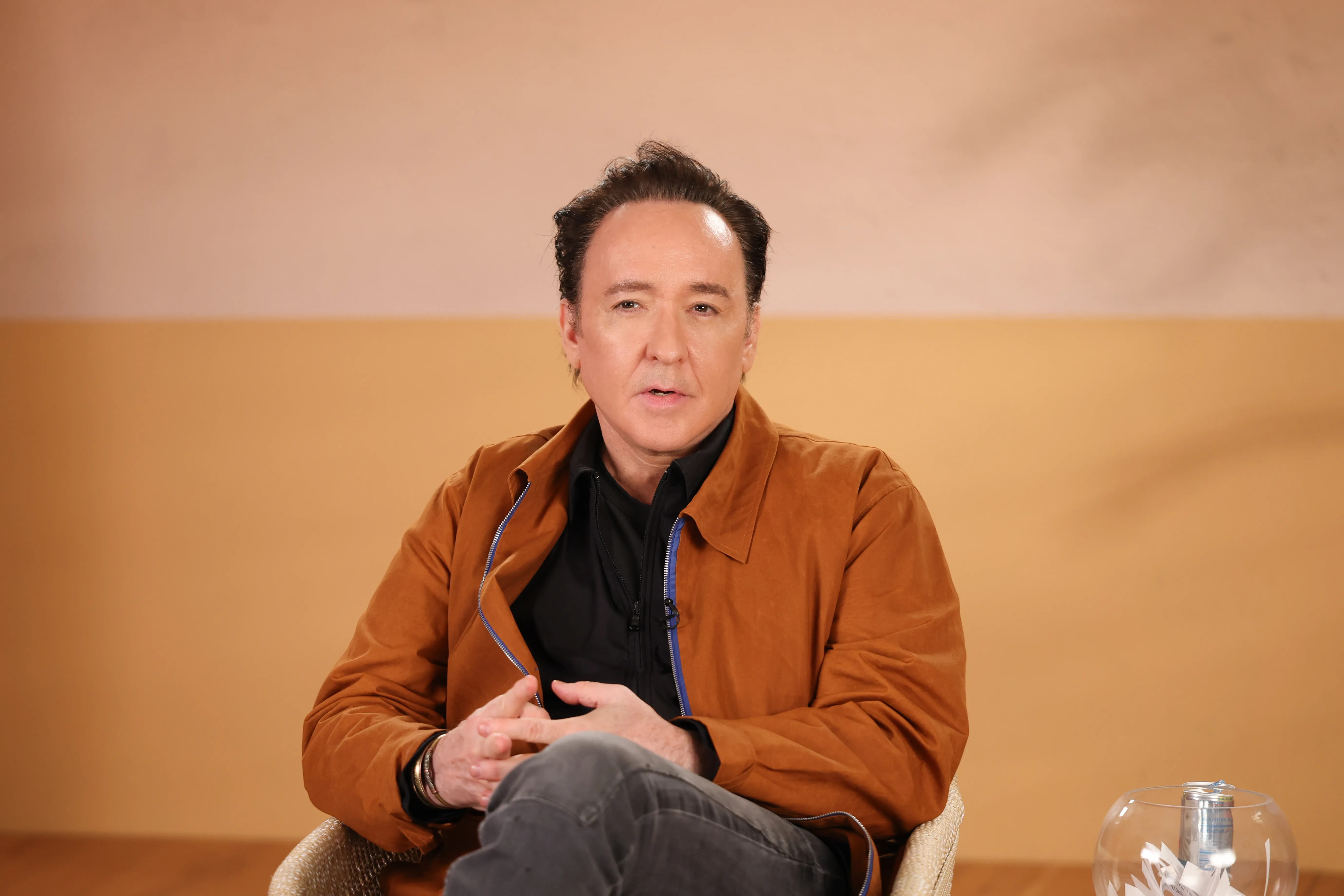 John Cusack seated and wearing a brown jacket over a black shirt.