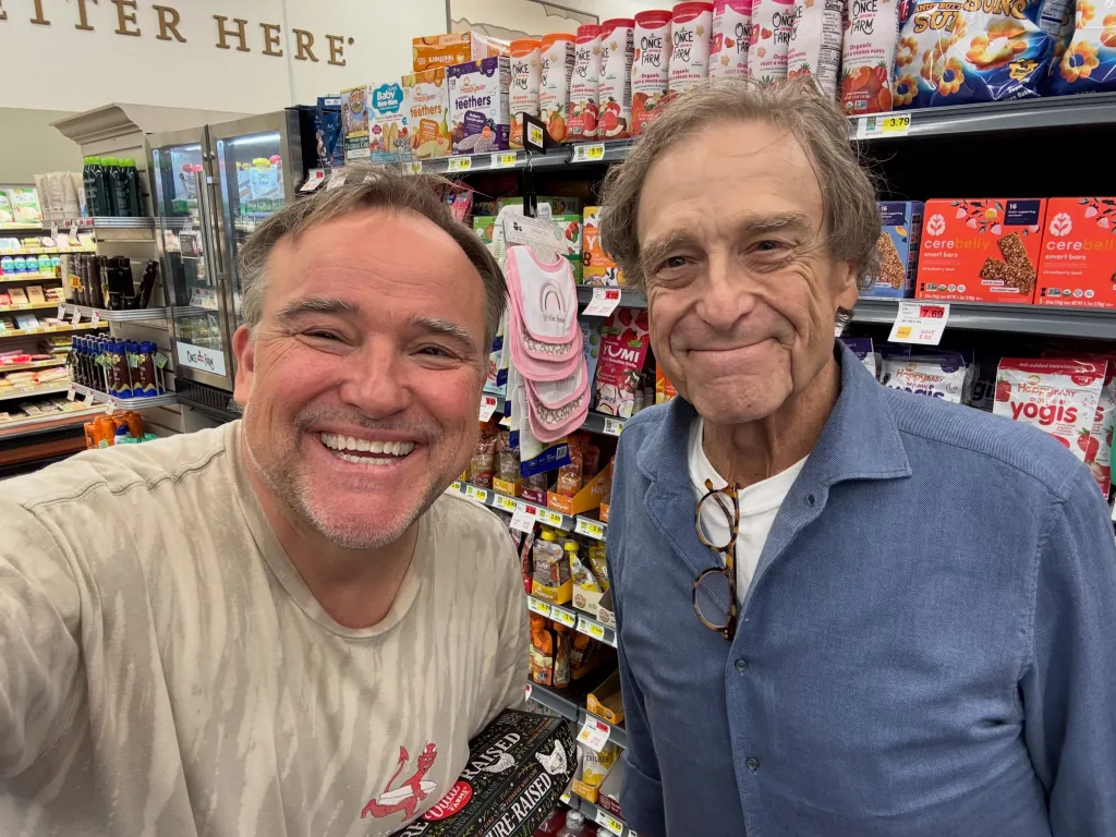 A man in a tan shirt smiles for a selfie with John Goodman, who is smiling, in a grocery store aisle.