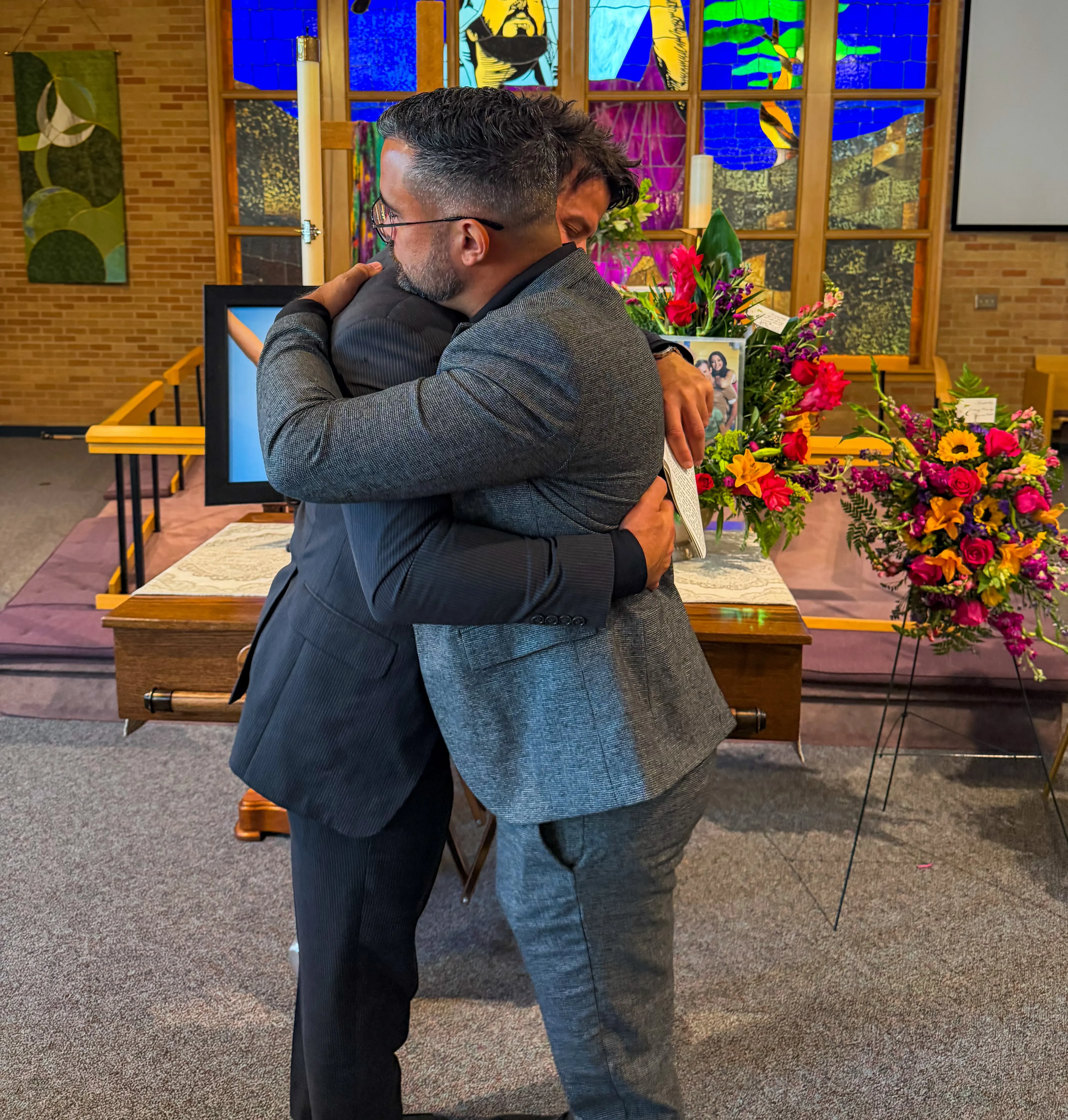 Two men embracing at a funeral service, with a casket and floral arrangements in the background.
