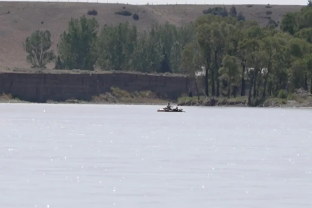 A person on a raft in the middle of a river with wooded banks and a distant hill.
