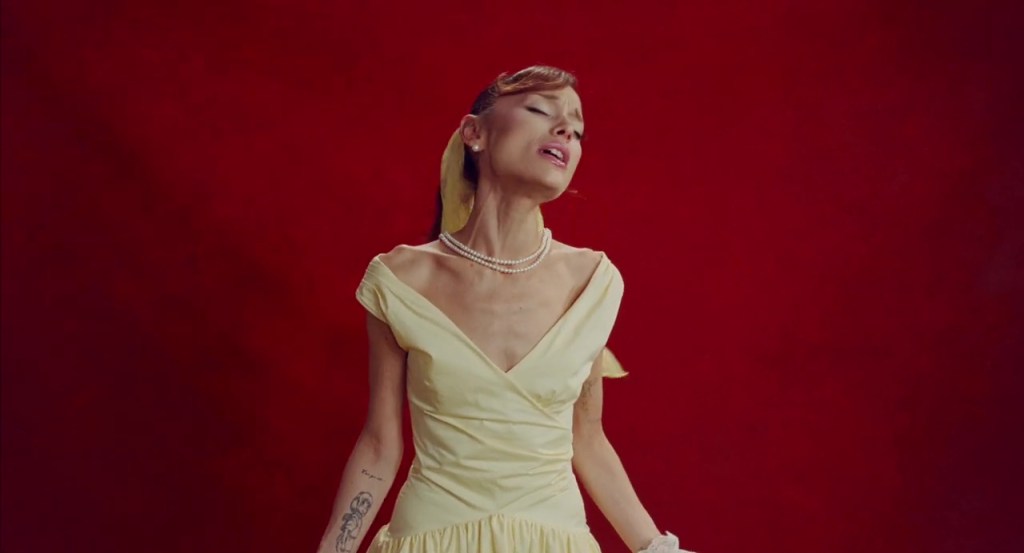 Ariana Grande singing in a yellow dress, with a yellow scarf and pearl necklace.