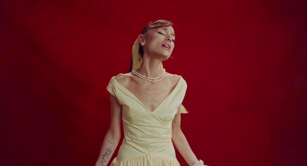 Ariana Grande singing in a yellow dress and pearls against a red background.