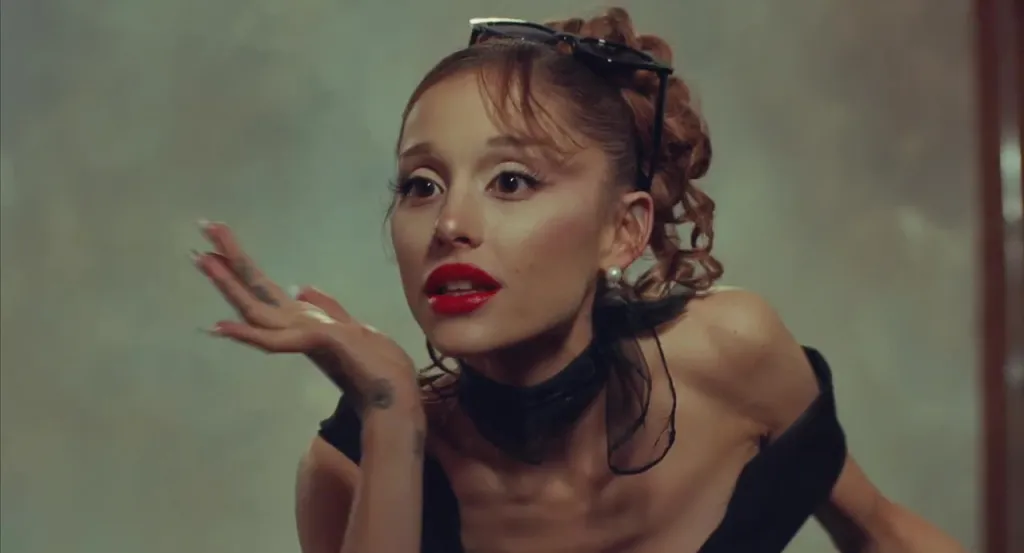 Ariana Grande in a black dress with red lipstick and sunglasses on her head, blowing a kiss.