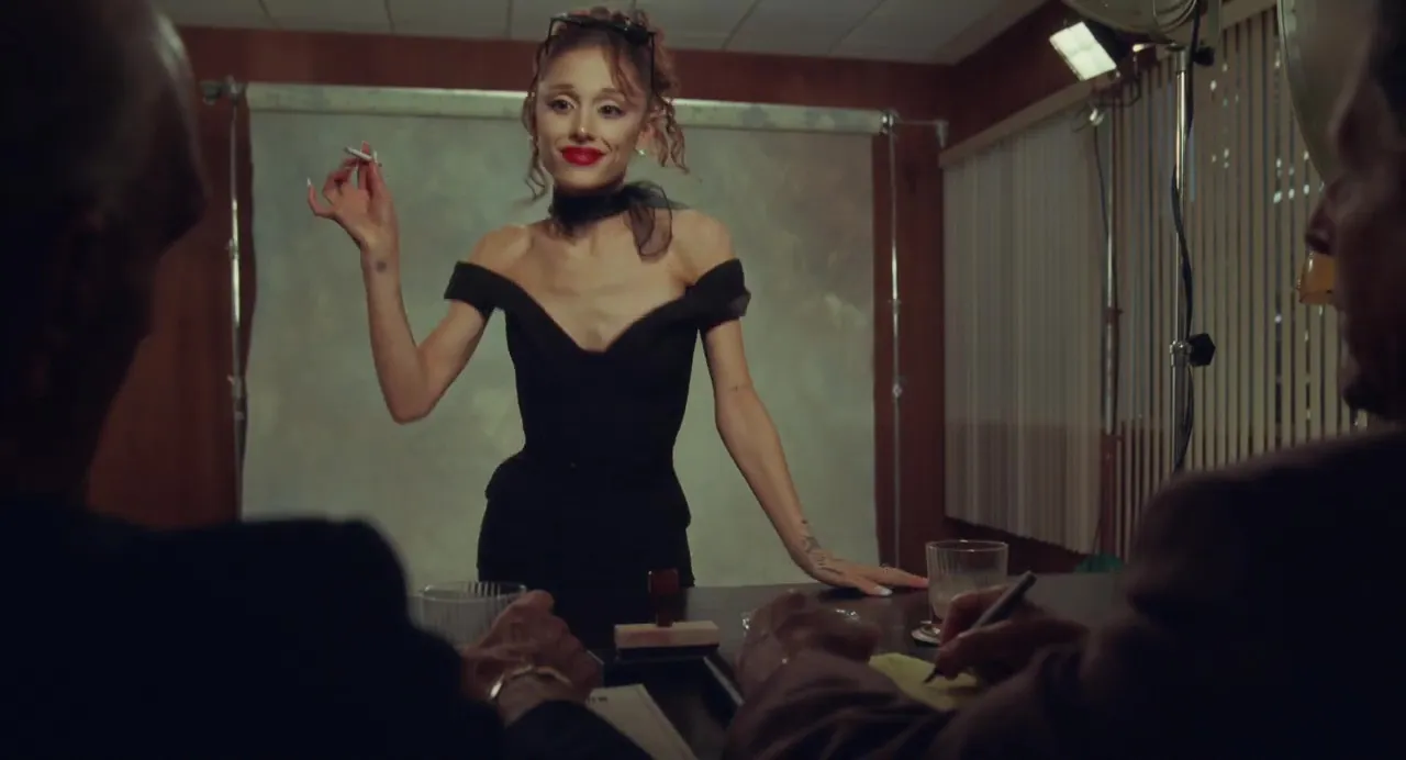 Ariana Grande in a black off-the-shoulder dress, red lipstick, and a black choker, holding a lit cigarette while standing behind a table with two men across from her.