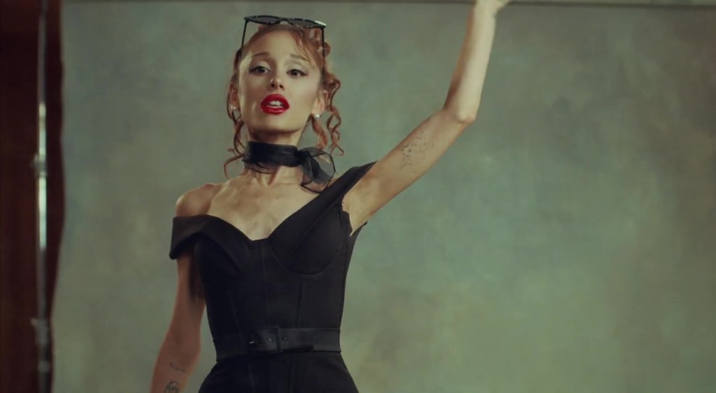Ariana Grande in a black off-the-shoulder dress with red lipstick, black neck scarf, and sunglasses on her head, with one arm raised.