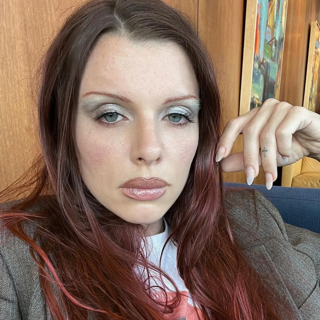 Julia Fox with red hair, frosted eyeshadow, and a cross tattoo on her finger.