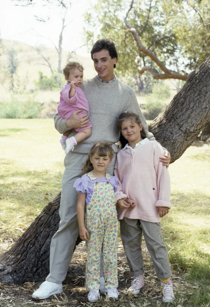 Bob Saget holding Mary-Kate or Ashley Olsen, standing with Jodie Sweetin and Candace Cameron.