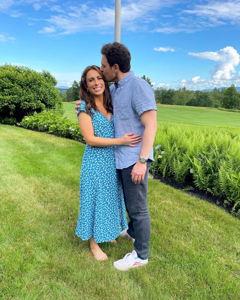 A man in a blue shirt kisses a woman in a blue floral dress on the forehead in a grassy outdoor setting.