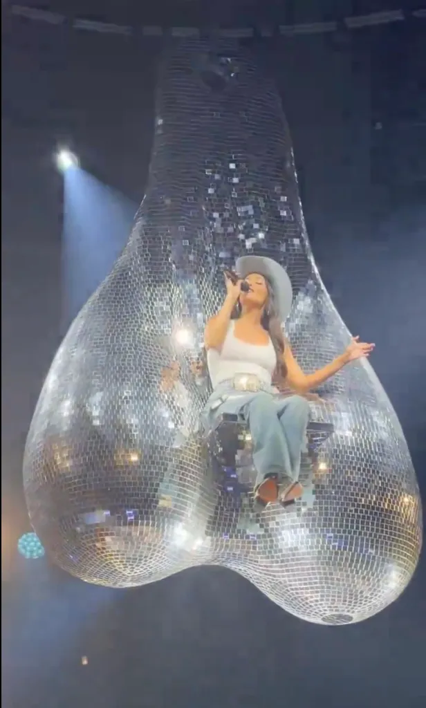 Kacey Musgraves in a cowboy hat and white tank top, sitting in a chair suspended within a giant disco ball structure on stage.