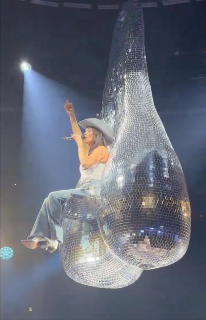 Kacey Musgraves in a cowboy hat and jeans straddling two large, reflective disco balls shaped like a pair of testicles during a performance.