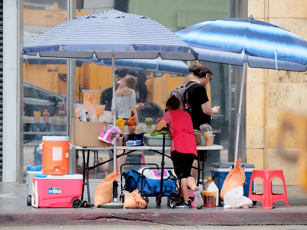 Two women operate a food stand on Hollywood Blvd under two umbrellas, selling fruit, drinks, and snacks.