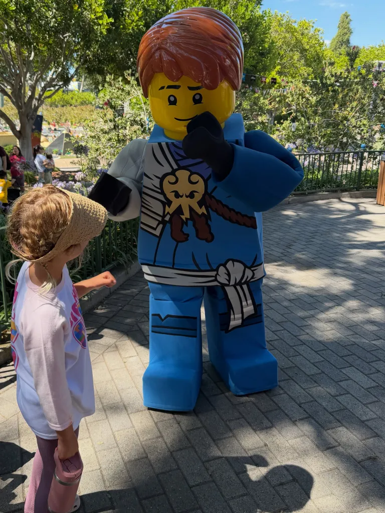 A child with blonde pigtails in a straw hat and a Lego character in a blue costume with orange hair standing on a brick pathway.