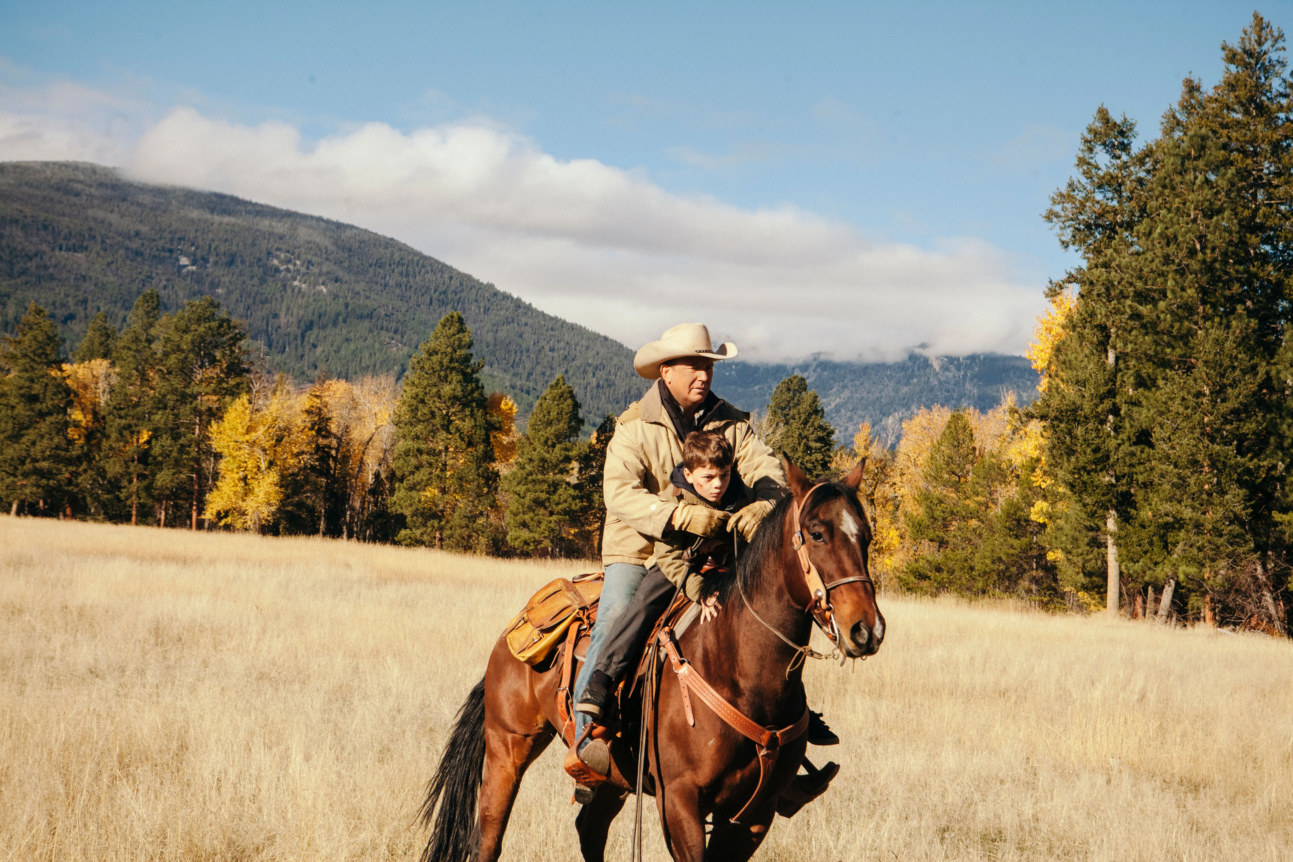 Kevin Costner and Brecken Merrill riding a horse in a field.