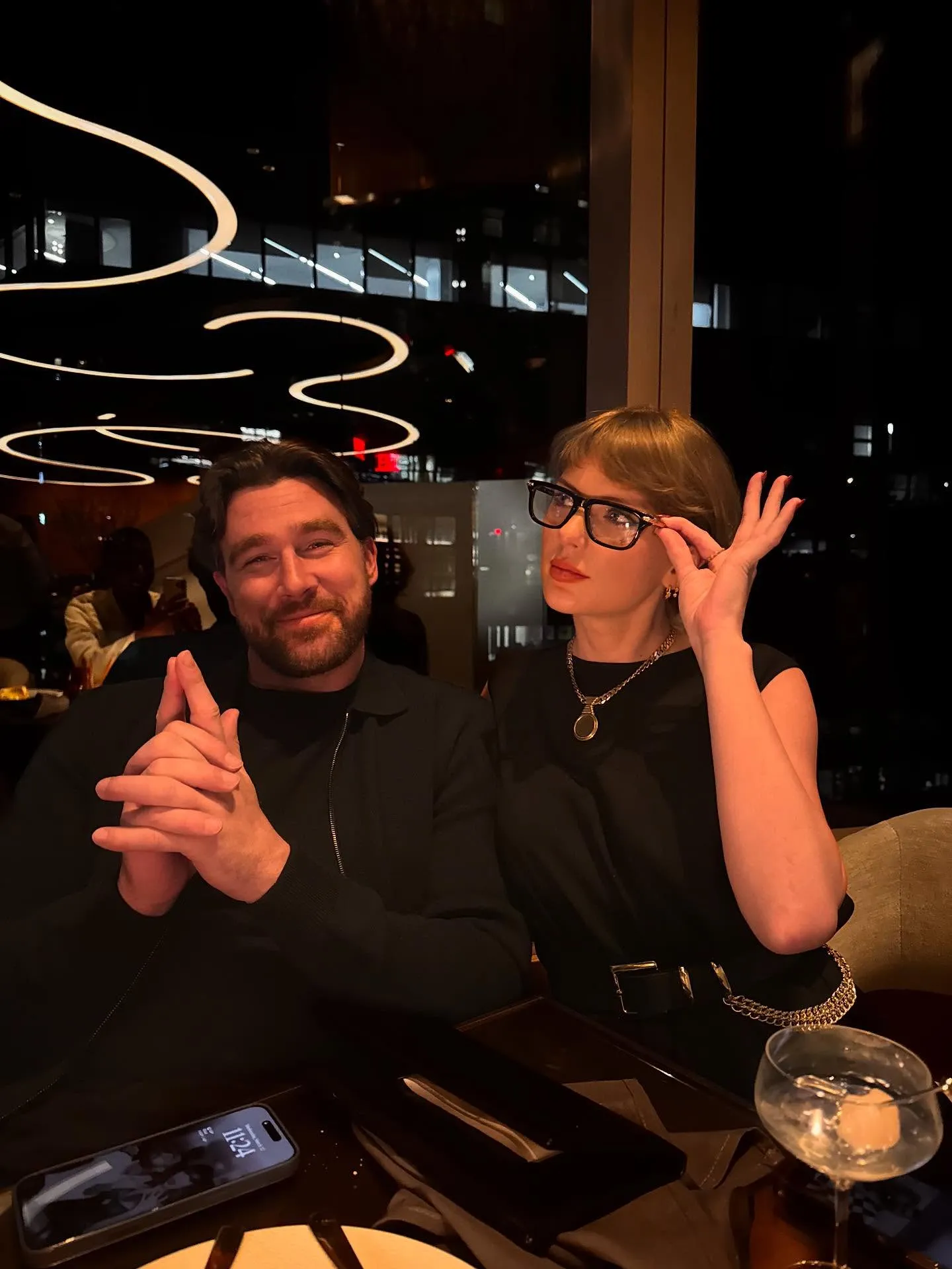 A man and woman at a restaurant, the man smiling, the woman adjusting her glasses.