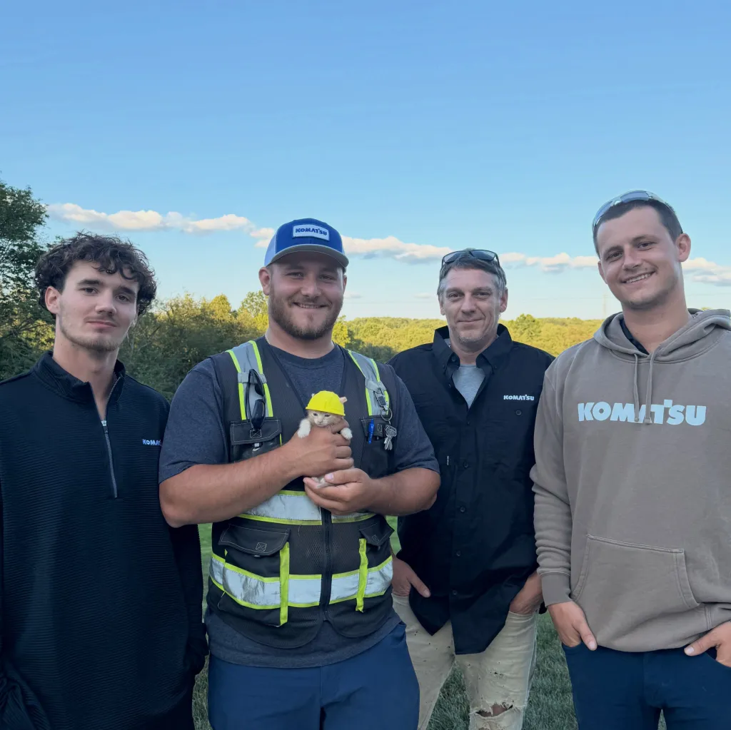 Four men and a kitten wearing a construction hat, with three men wearing Komatsu-branded apparel.