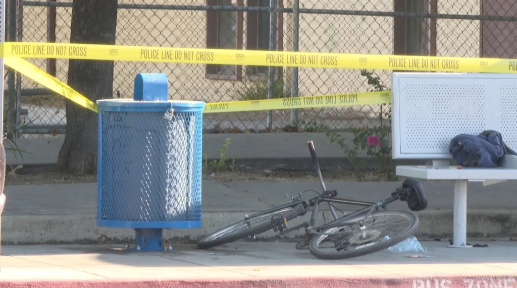 A crime scene with police tape, a bicycle, a blue trash can, and a bench with clothes.