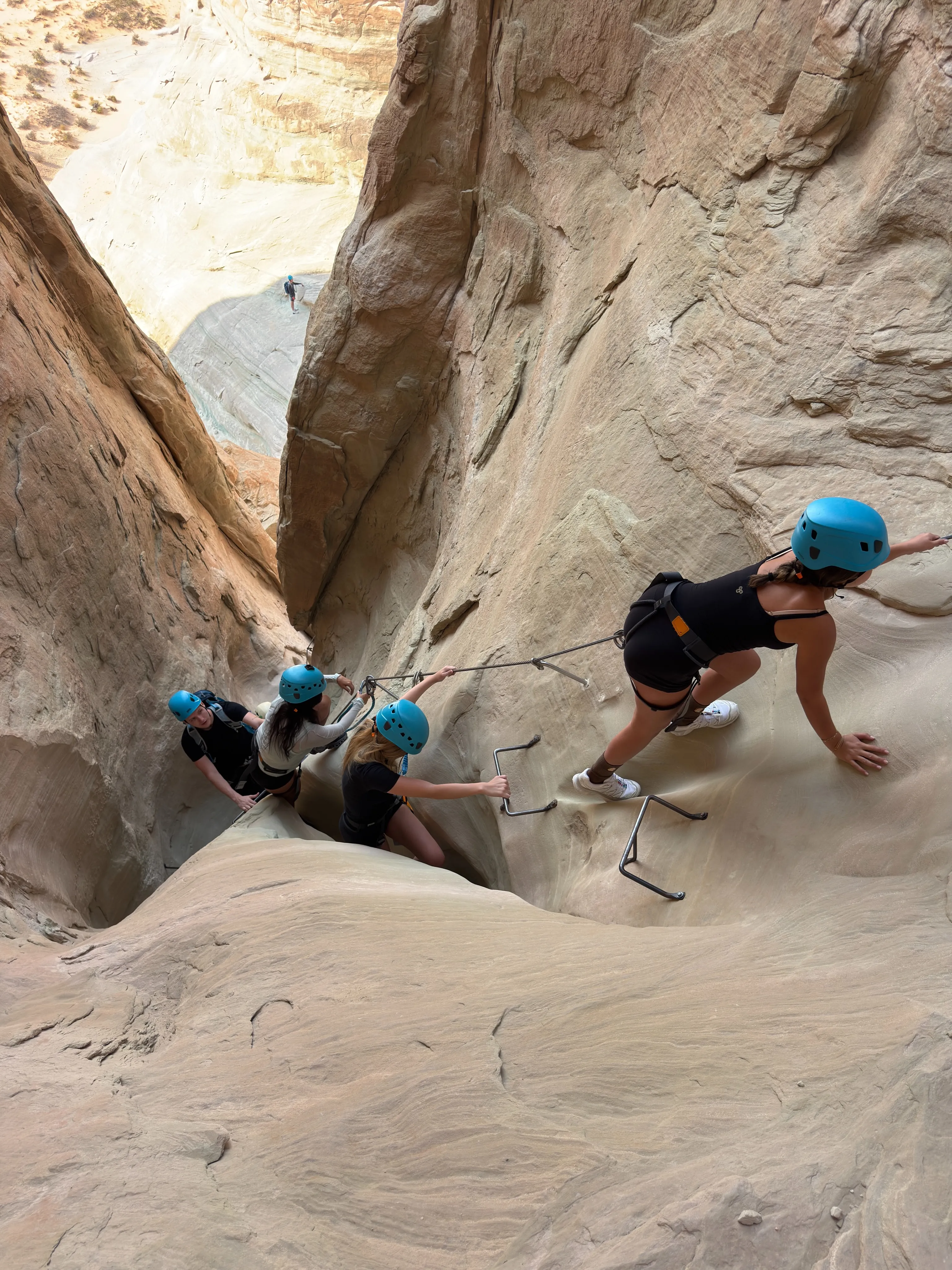 People in helmets and harnesses climbing a steep rock face with attached ropes and foot holds.