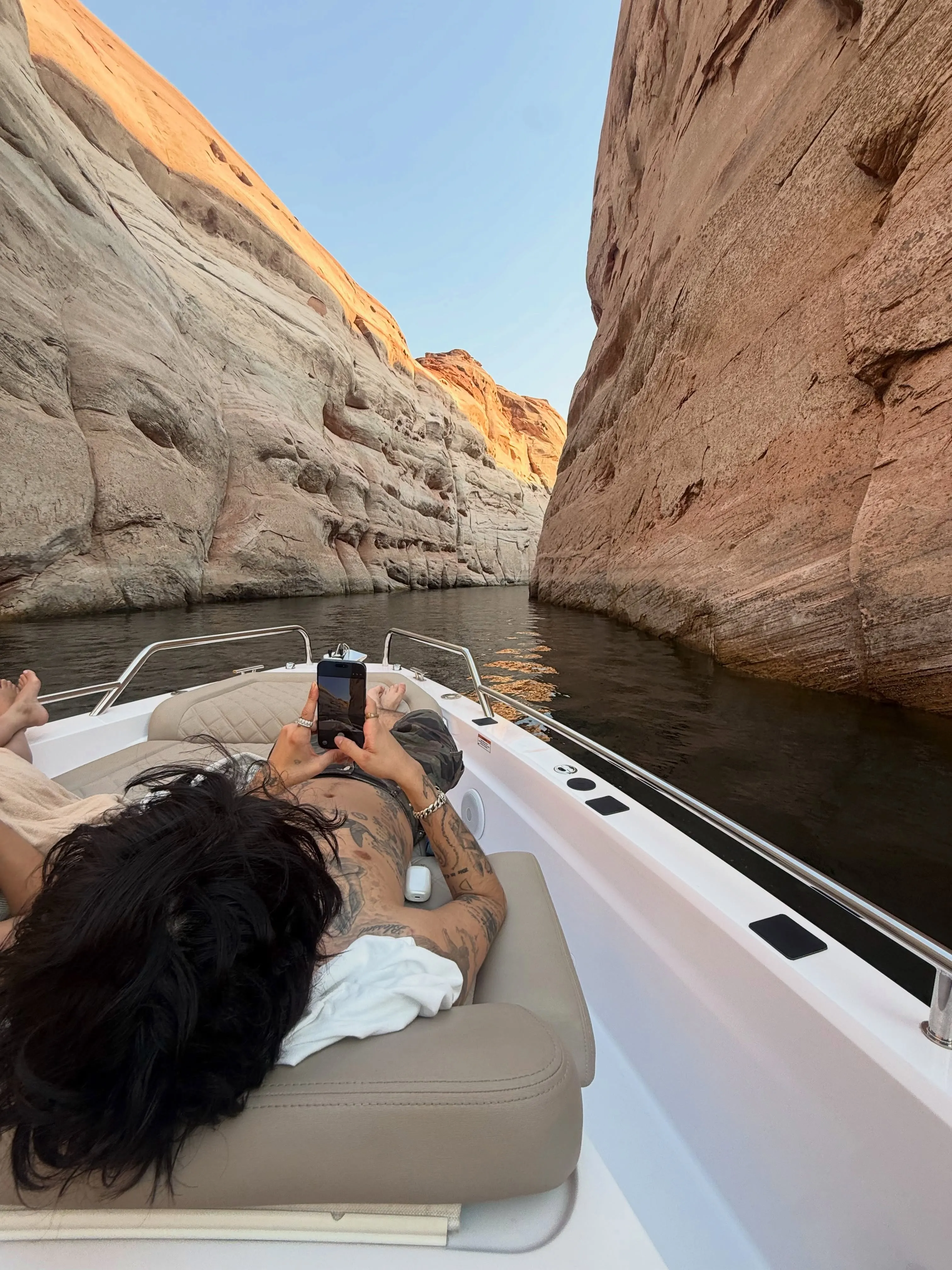 A person with tattoos on their chest and arms lies on a boat, holding a phone to take a photo of the canyon walls rising from the water.