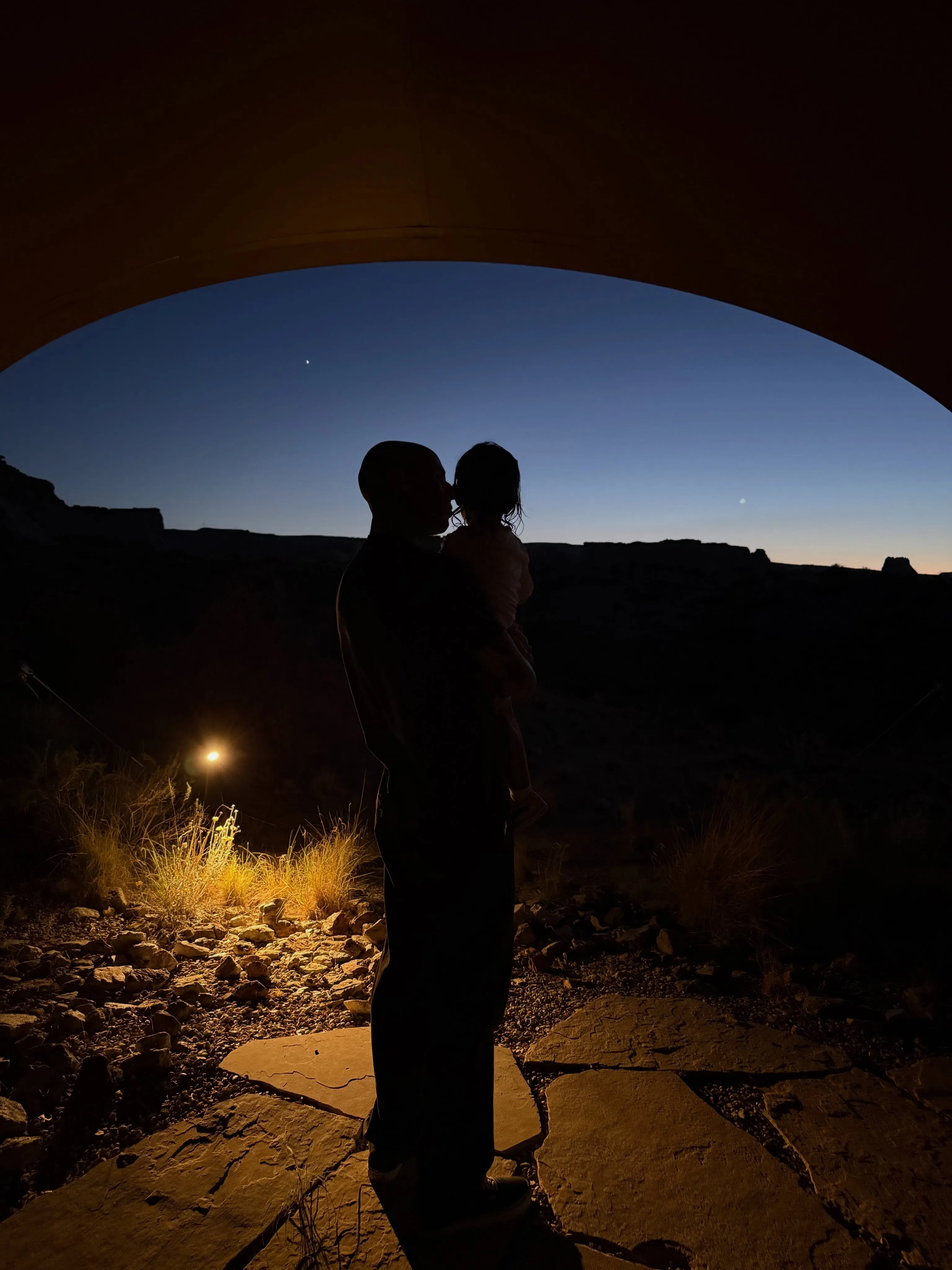 Silhouette of a Travis Barker holding a baby against a dark blue twilight sky with mountains in the background.