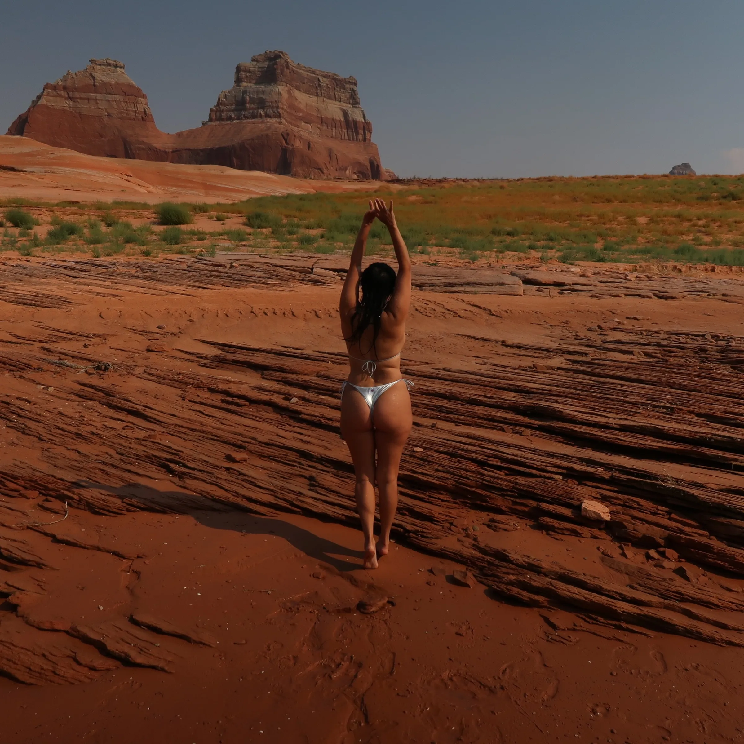 Kourtney Kardashian in a white thong bikini with her arms raised, standing on red rock formations.