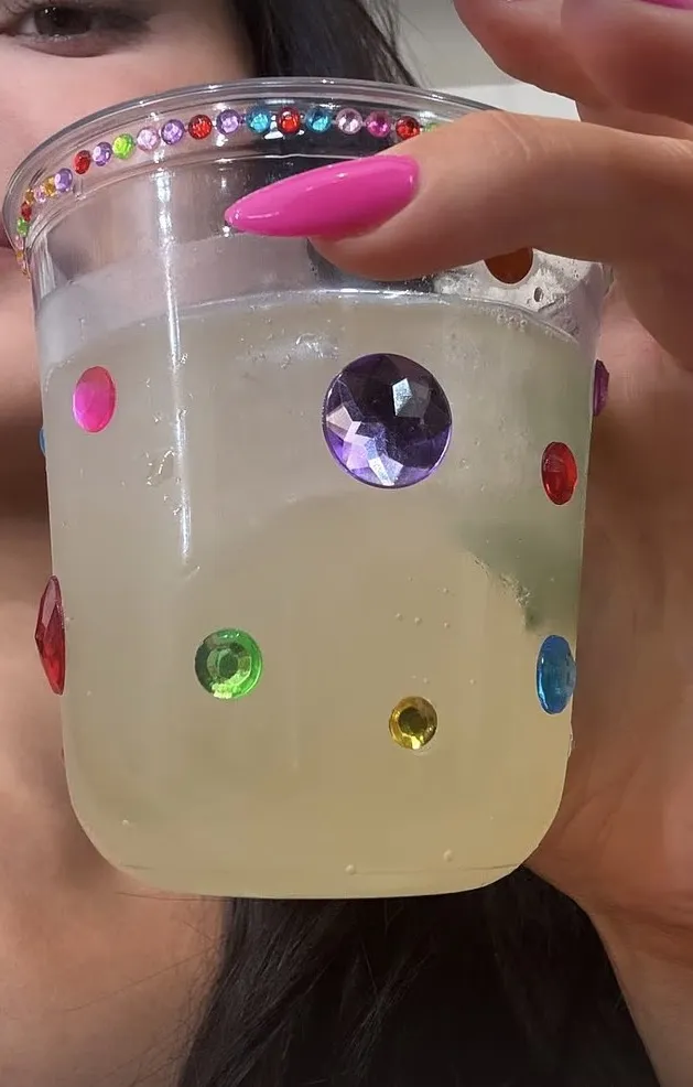 A person holding a clear plastic cup decorated with colorful gems, filled with a light-colored beverage.
