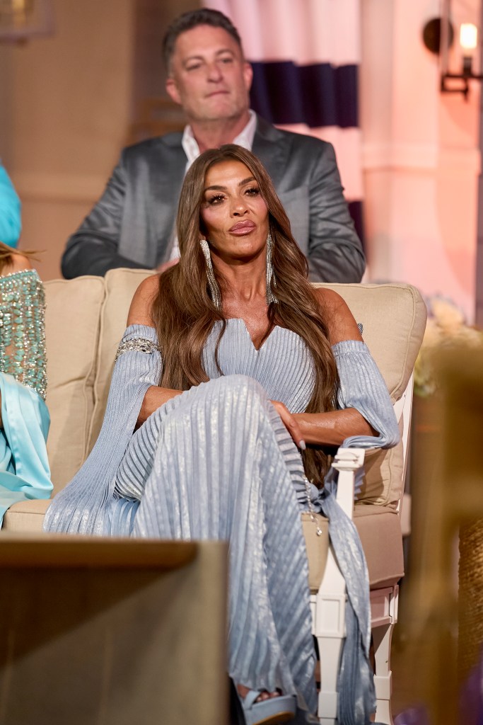 Liz McGraw in a light blue off-the-shoulder pleated dress with long earrings, sitting in a beige armchair.