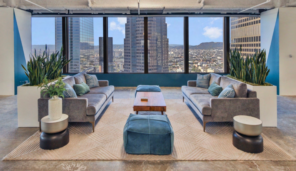 A modern office lounge with two grey sofas facing each other, separated by a wooden coffee table and a blue ottoman, with large windows overlooking a city skyline.