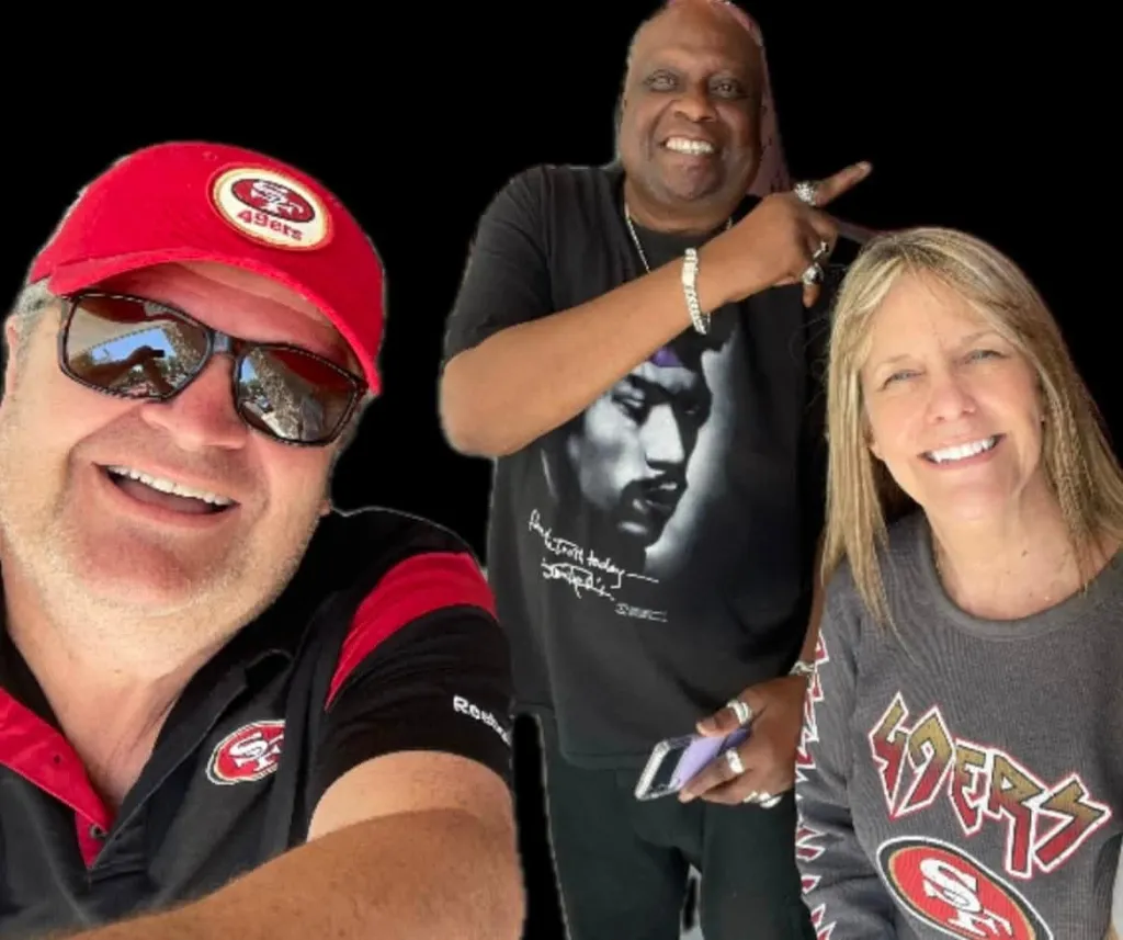 Lamont Hollywood, a man in a black T-shirt with a picture of Tupac, smiles and points with a woman and another man in a red hat.