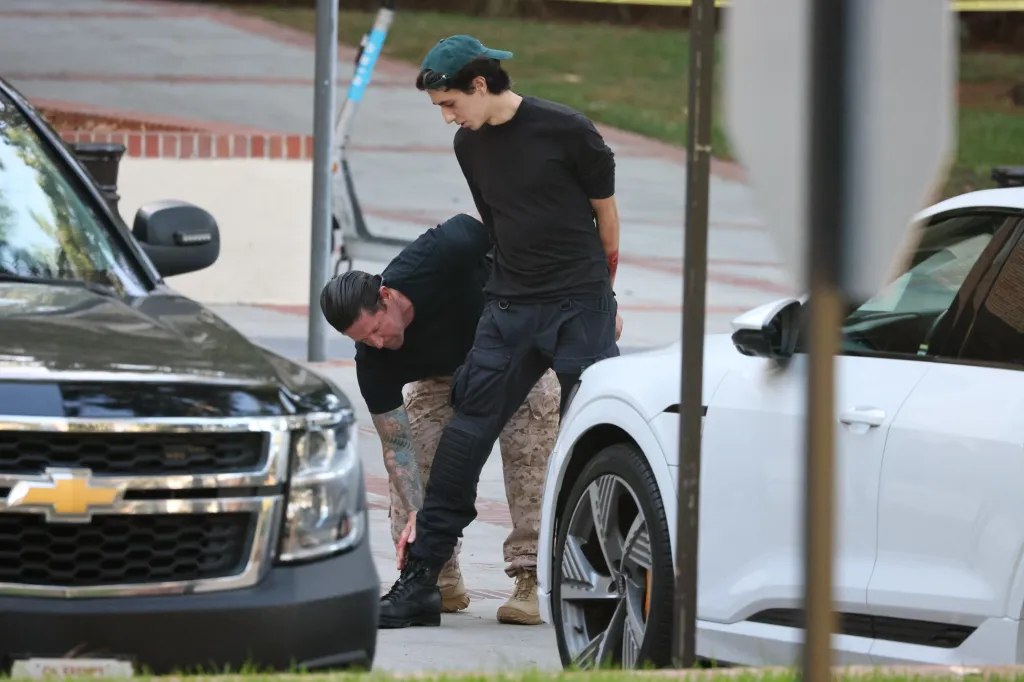 A man in a black shirt and camouflage pants bending down, adjusting the boot of another man in a black shirt, with a red injury on his arm, standing between a black and a white car.