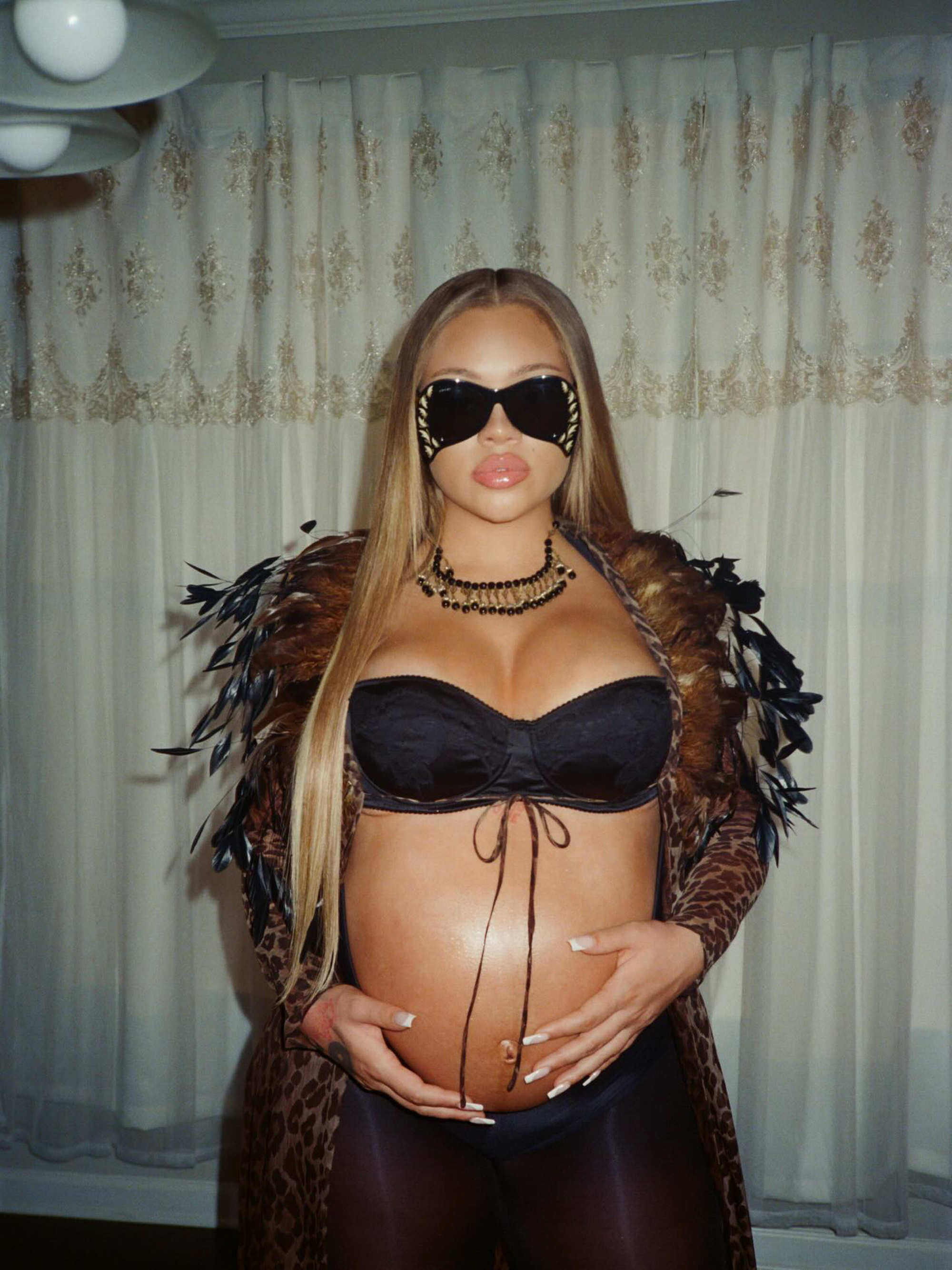 Latto cradling her pregnant belly while wearing a black bra, sheer leopard print and feathered robe, black sunglasses, and black and gold necklace.