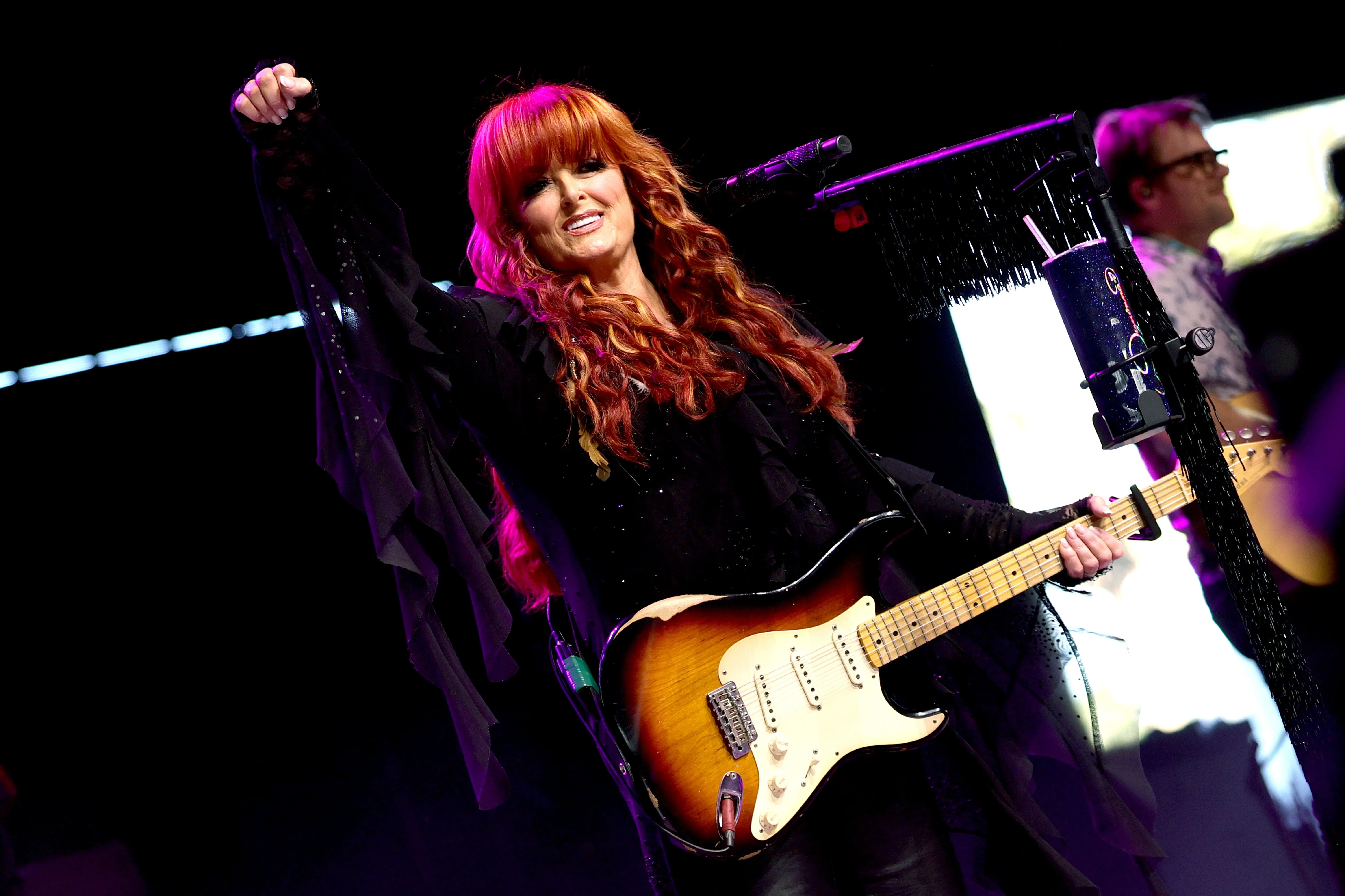 Wynonna Judd performing on stage at the 2026 Stagecoach Festival with a guitar, wearing a black shirt with ruffles, and raising her fist in the air.