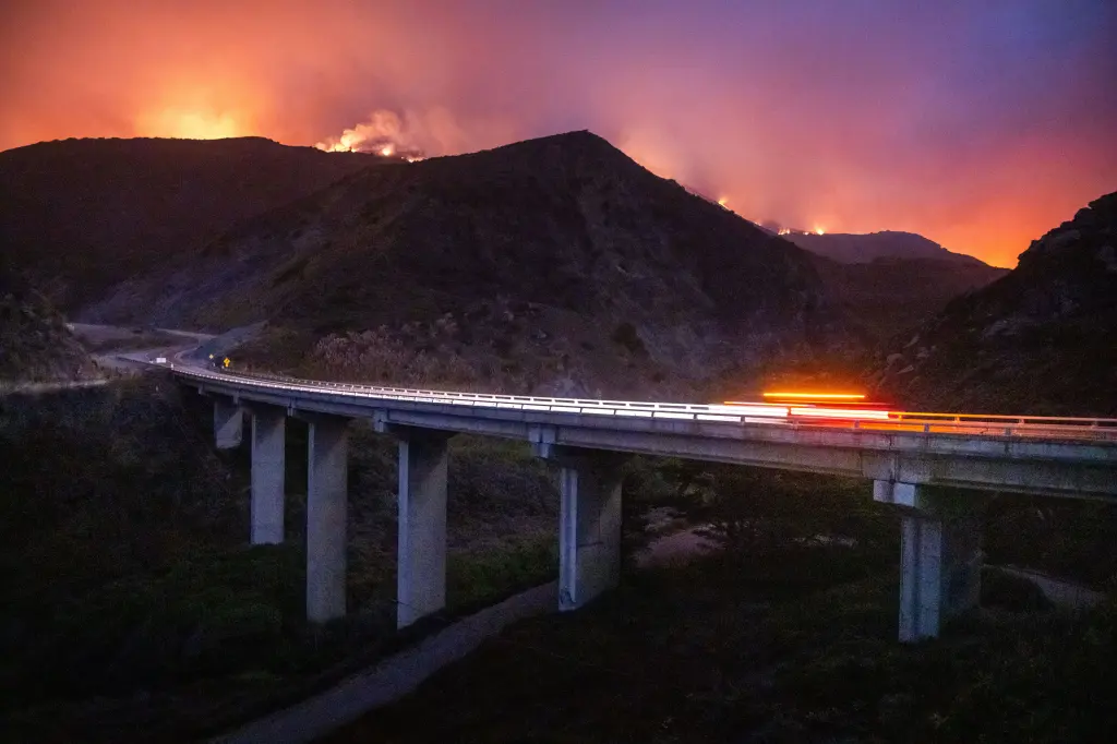 A highway bridge with car lights streaks across a dark landscape, with hills burning in the background under an orange and purple sky.