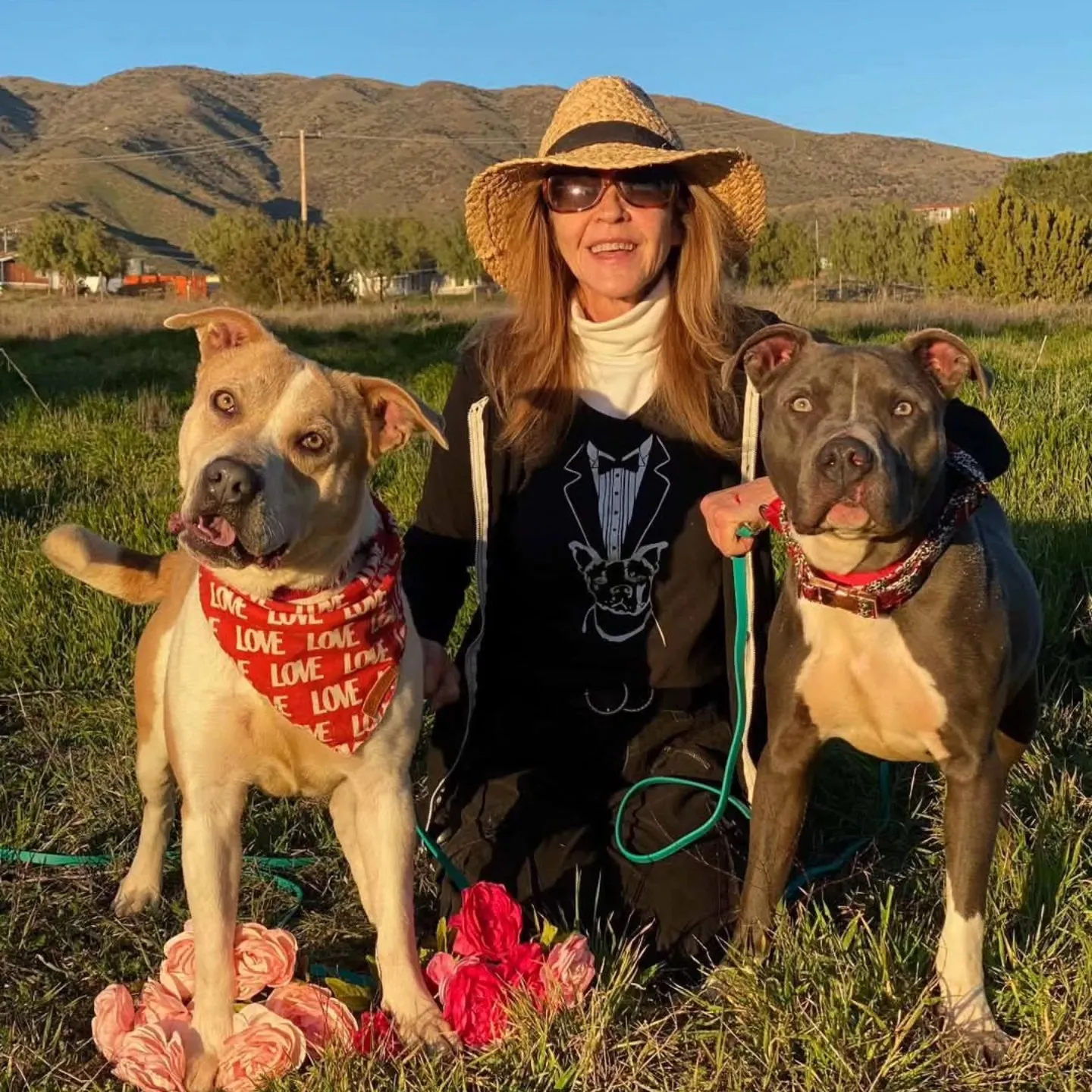 Linda Blair kneeling with two dogs in a field with mountains in the background.