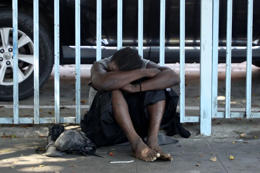 A homeless person sits on the ground with their head in their arms, with a fence and a car tire in the background.