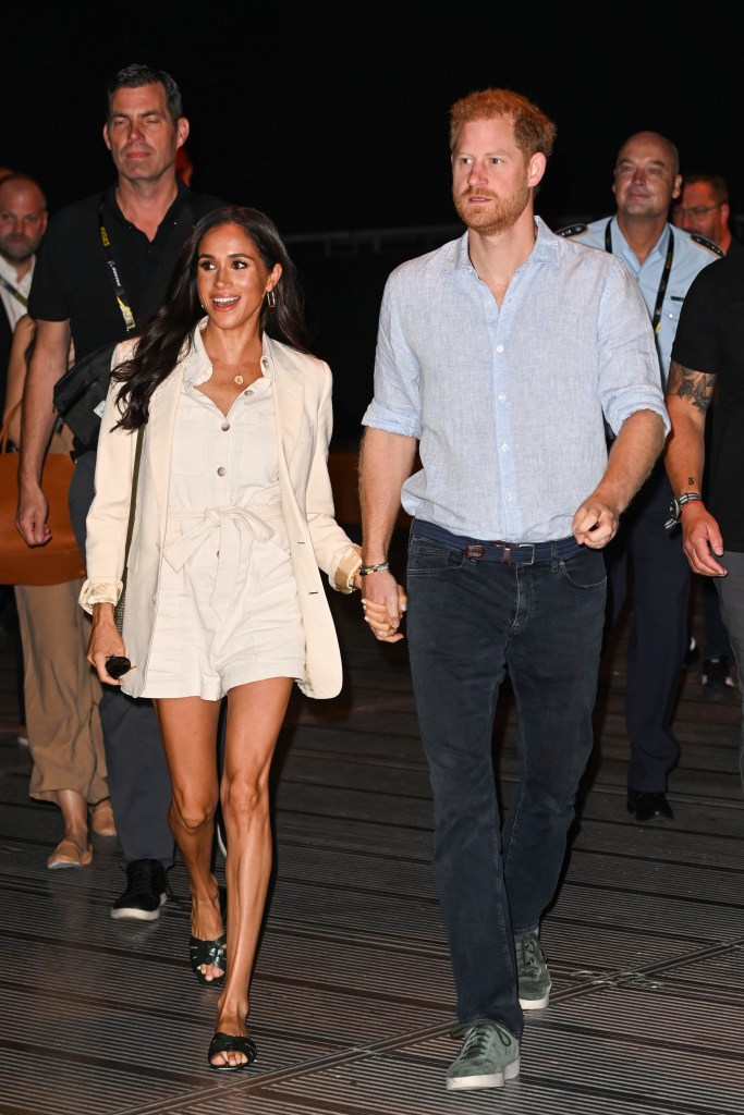 Meghan Markle, in a white shirt and white short and cream jacket, and Prince Harry, in a blue shirt and black jeans, holding hands while walking, with security personnel in the background.