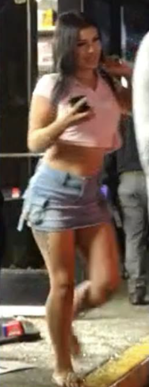 Suspect with long dark hair, wearing a pink crop top and a denim mini skirt, holding a phone.