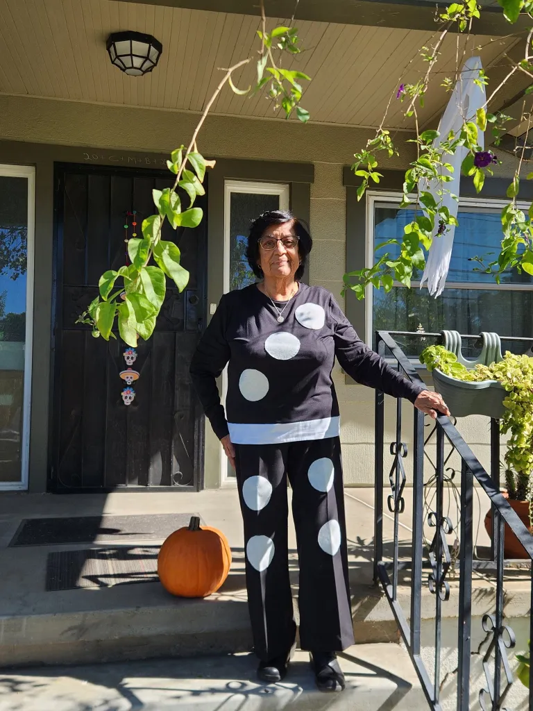 Cristina Vargas, wearing a black outfit with white polka dots, stands on her porch with a pumpkin nearby.