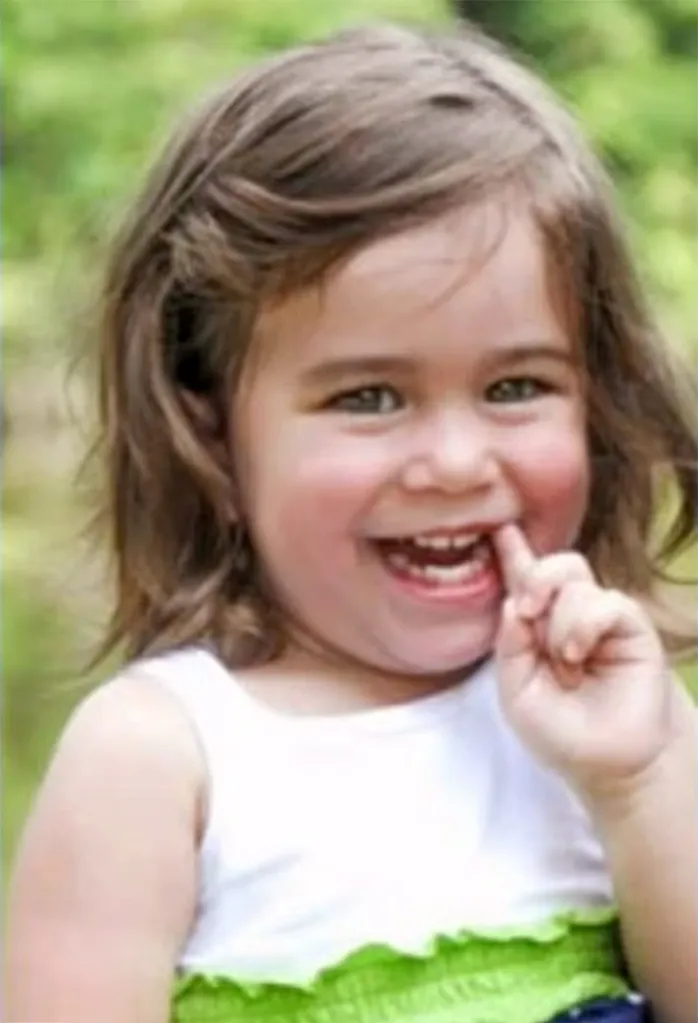 Macy Grace Ditty, a 2-year-old girl with brown hair, smiling with her finger near her mouth.