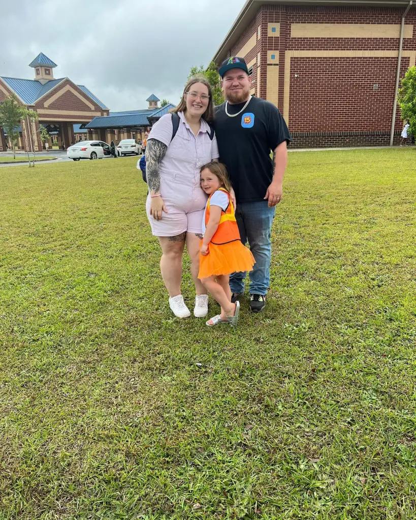 A woman in a light pink jumpsuit, a man in a black t-shirt and jeans, and a young girl in a white shirt with an orange vest and tutu stand in front of a school building.