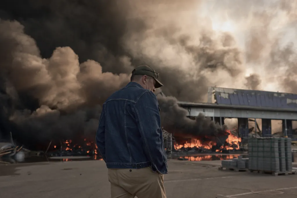 A man in a baseball cap and denim jacket looks on as a warehouse burns, with large plumes of black smoke rising into the sky.
