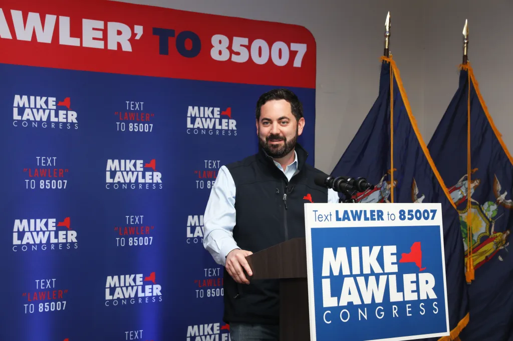 Rep. Mike Lawler stands at a podium in front of a blue backdrop with his campaign logo and instructions to text 