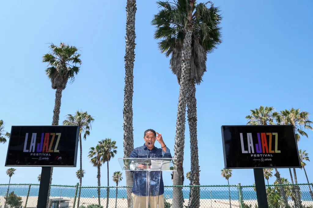 Martin Ludlow, CEO and founder of the Los Angeles Jazz Festival, speaks at a podium with two large screens displaying the 