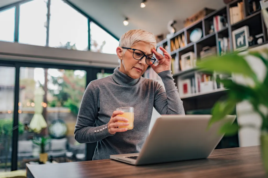 A mature woman holds her head while looking at a laptop, experiencing a headache.
